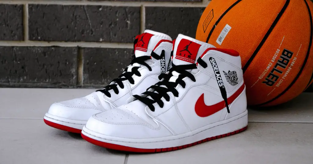 Photo of two white Jordan basketball shoes with black laces and red Nike swoosh, side by side on the floor, a basketbal behind them.
