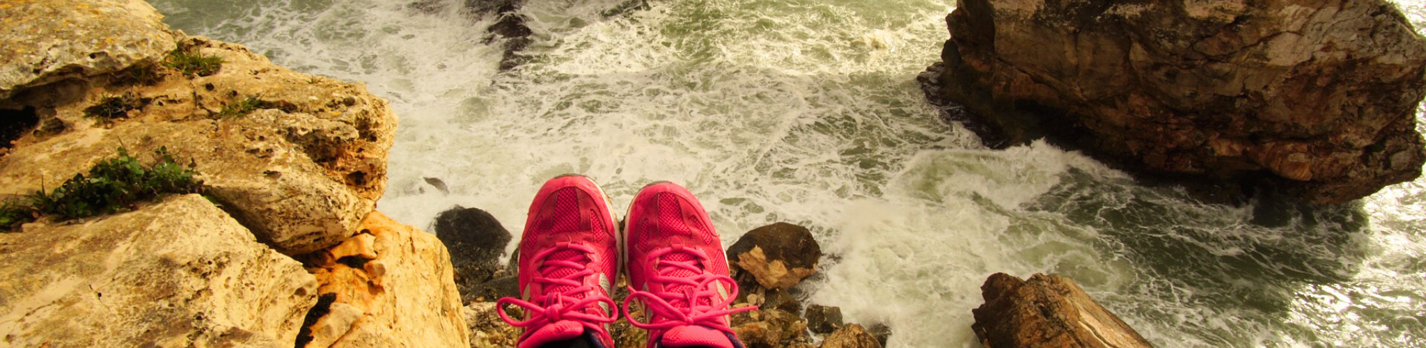 Stock photo of someone sitting on the edge of a rock over the ocean, wearing pink hiking sneakers.