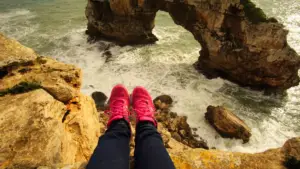 Stock photo of someone sitting on the edge of a rock over the ocean, wearing pink hiking sneakers.