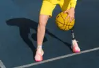 Stock photo of person from the thighs down, wearing yellow shorts and pink basketball shoes, dribbling a basketball on a blue basketball court outdoors. The leg on the right side is a prosthetic leg.