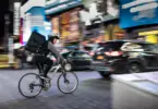 Stock photo of a person on a bike with a delivery backpack on, in a cityscape.