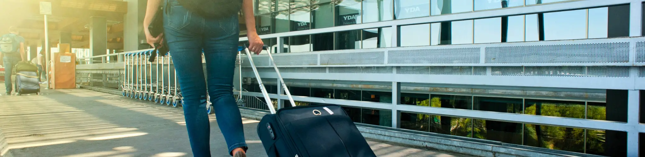 Stock photo of a person with a suitcase walking down a sidewalk by an airport.
