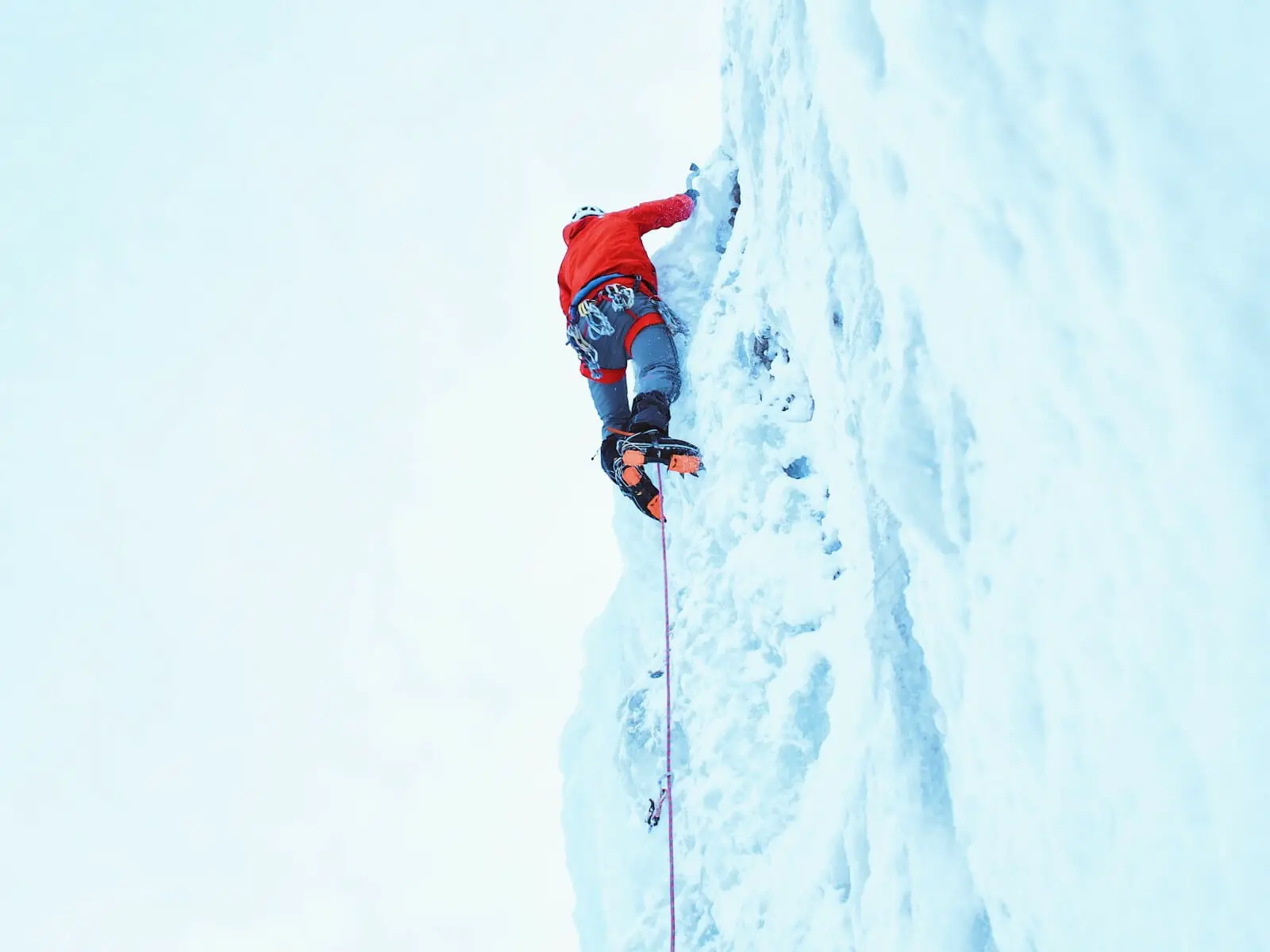 Ice climber in bright red jacket with 8-point crampons and safety rope climbing ice flow.