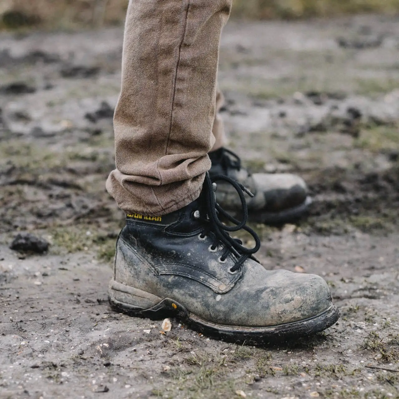 Person standing in muddy black hiking boots with oversized laces on a mud trail with sparse blades of grass.