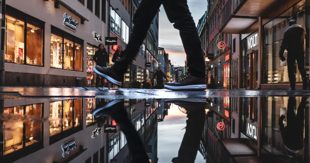 Stock photo of legs from the knees down, walking through a puddle in the middle of a shopping street with restaurants and bars on either side.