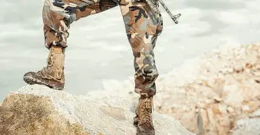 Person standing on rock in tactical boots with rifle and camouflage pants.