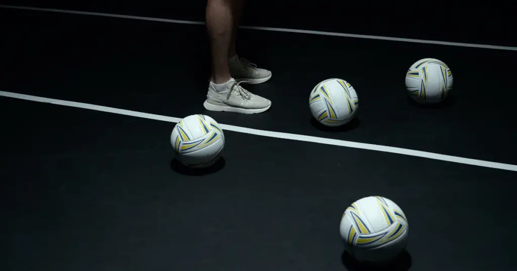 The legs of a person, top center, wearing the white and gold basketball shoes on a black court next to four white volleyballs.