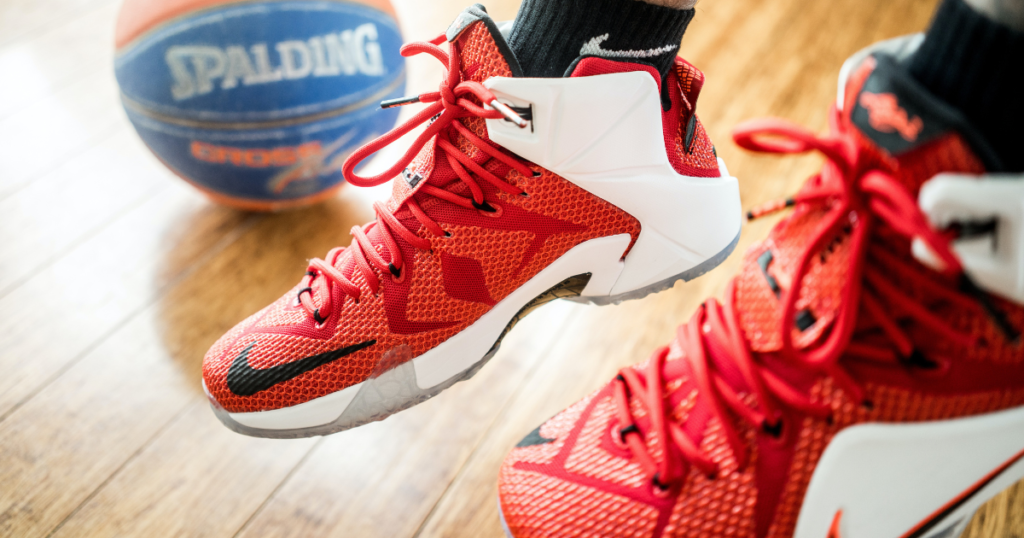 Closeup of someone's basketball shoes with a basketball and wooden court in the background. Foreground: bright red shoes with black Niken swoosh on toe, dark red design on sides, and white heel, sole, and upper sides.
