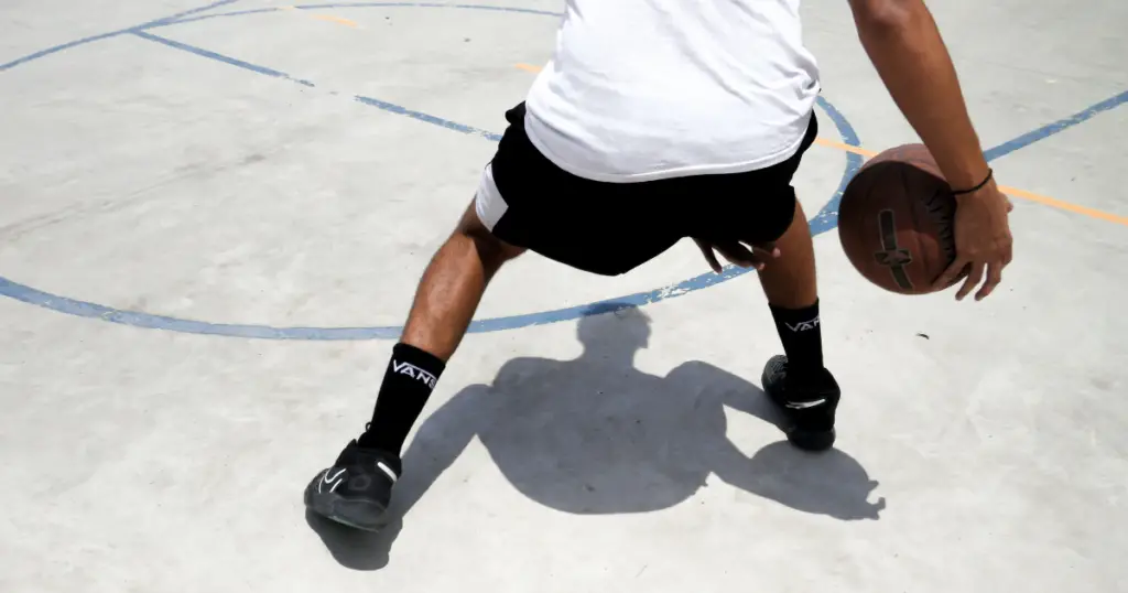 Photo from behind a basketball player in a white shirt and black shorts and black baskeball shoes, dribbling a basketball on a white court.